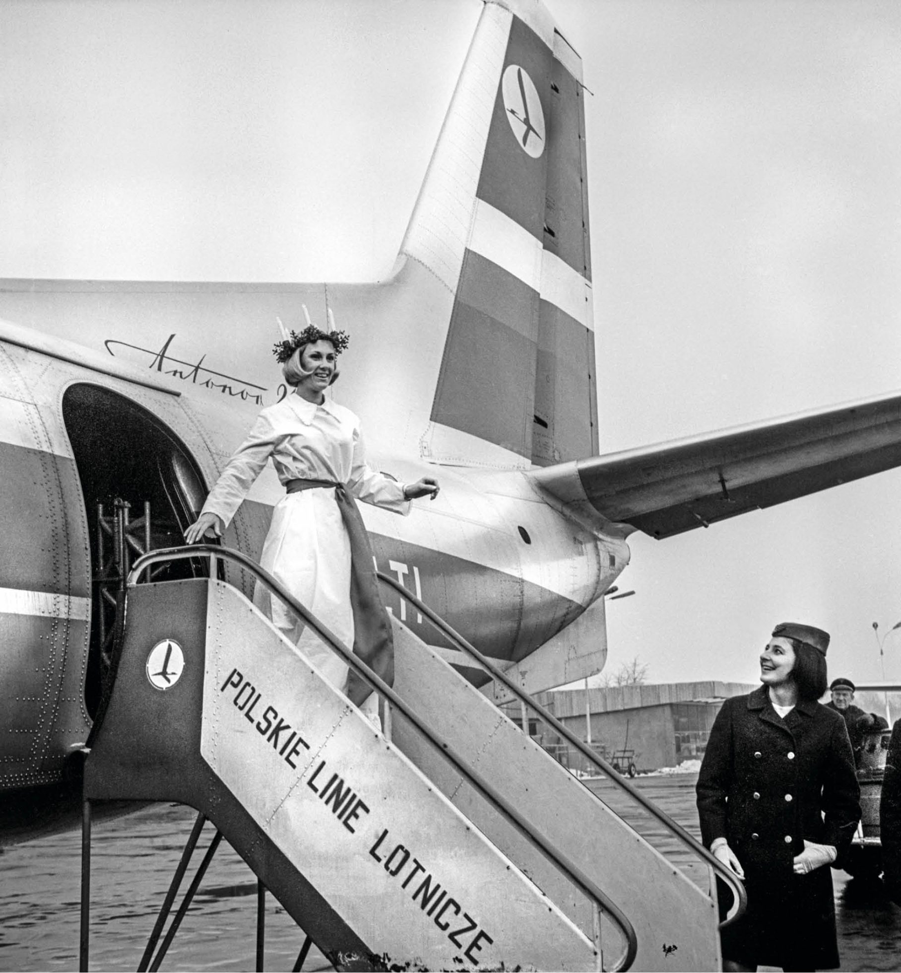 A black and white photo of a dressed-up woman with a wreath, getting off a plane.