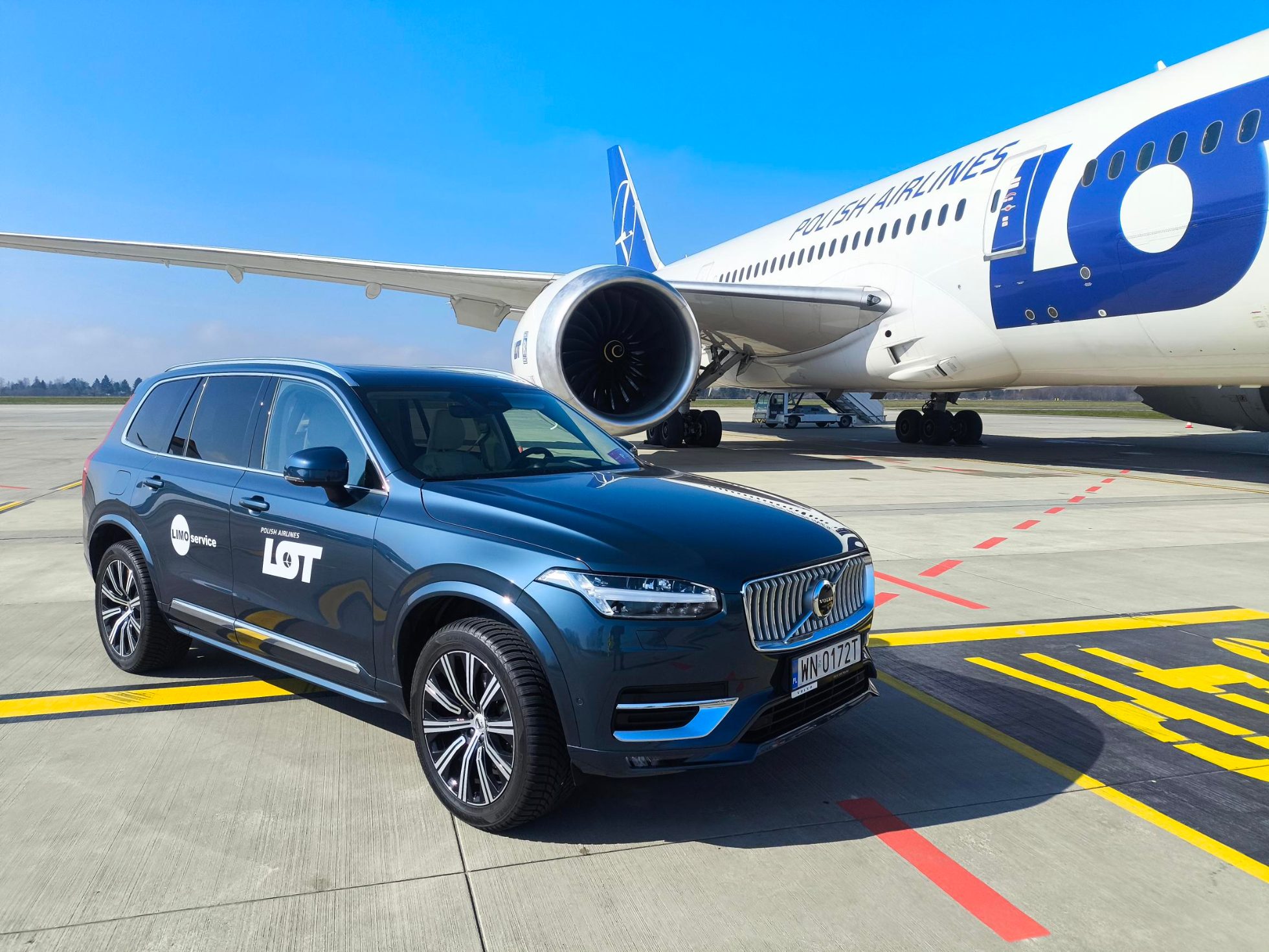 Luxury limousine awaits at the airport. Black paint gleams in the sun, driver opens the door.
