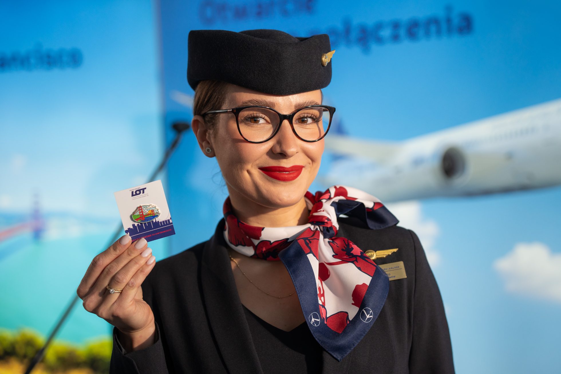 A LOT flight attendant in uniform, wearing a cap, holding a colourful LOT San Francisco pin