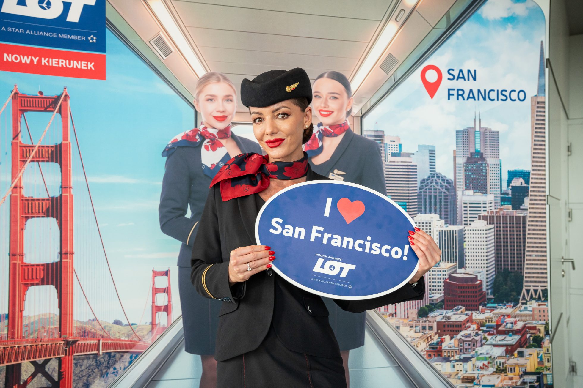 An LOT flight attendant in uniform holding a navy blue sign with the white words ‘I LOVE SAN FRANCISCO’ and the LOT logo