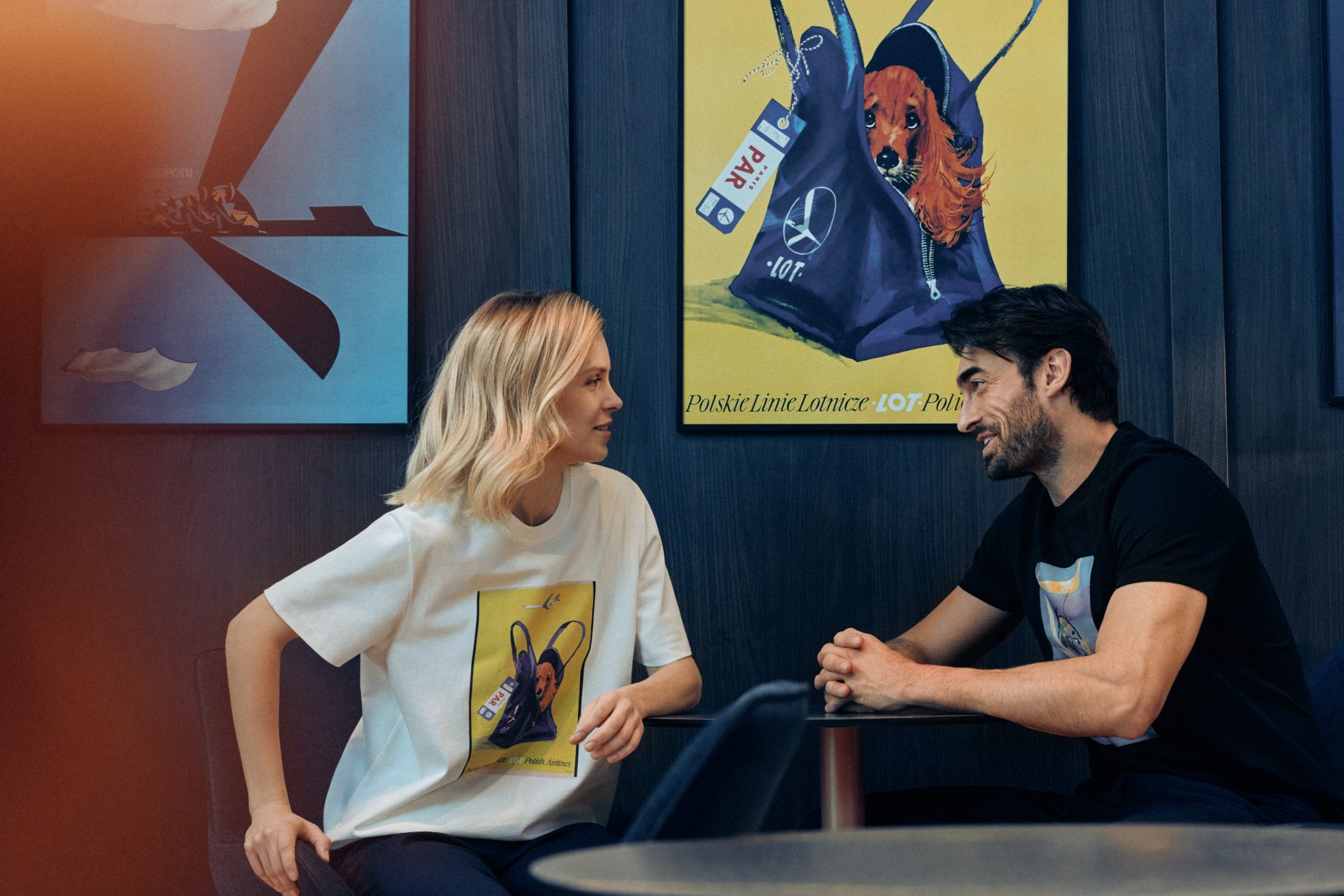 Mężczyzna i kobieta w koszulkach z nadrukami z kultowych plakatów LOT-u, siedzący i rozmawiający w LOT Business Lounge Mazurek
