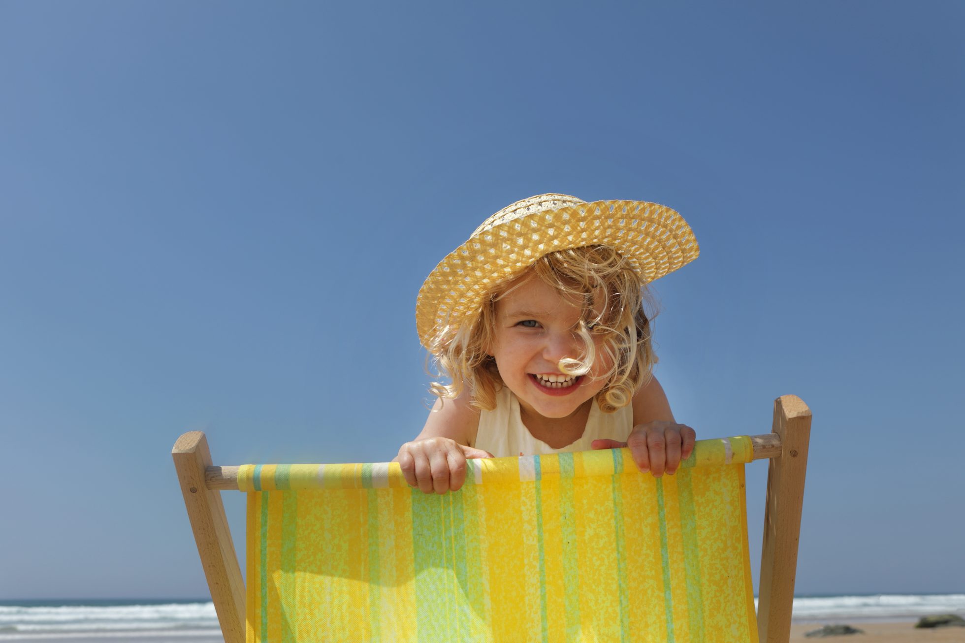 mała dziewczynka o blond włosach w kapeluszu opiera się o kolorowy leżak na plaży, zdjęcie na tle niebieskiego, czystego nieba