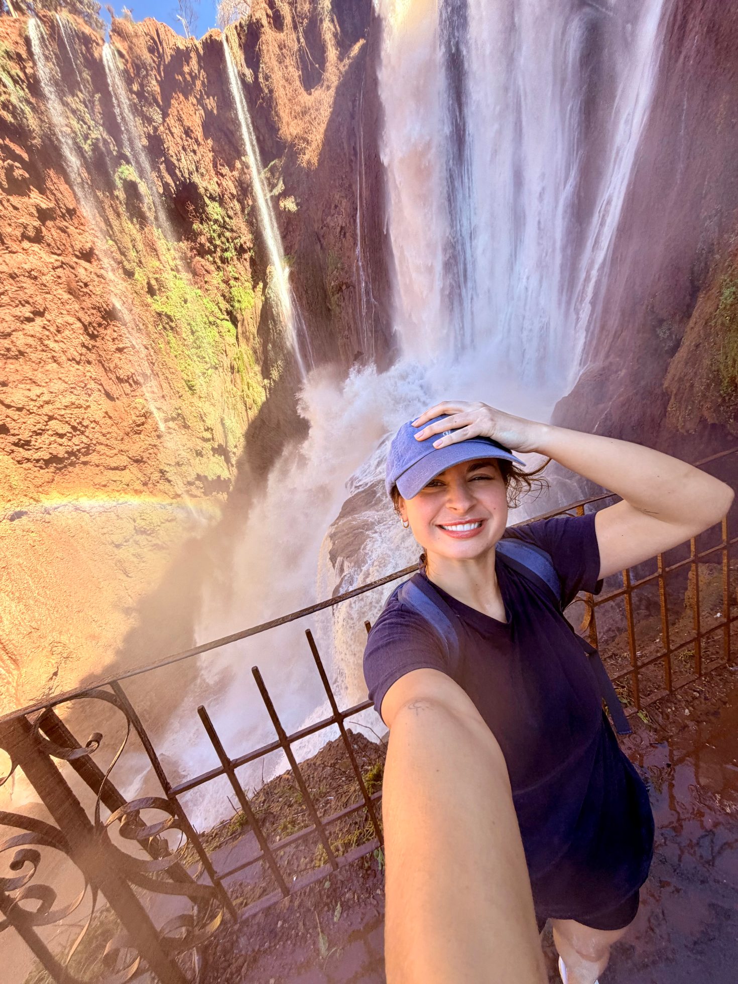 Uśmiechnięta kobieta w niebieskiej czapce z daszkiem robiąca sobie zdjęcie selfie przy barierce z widokiem na potężny wodospad.