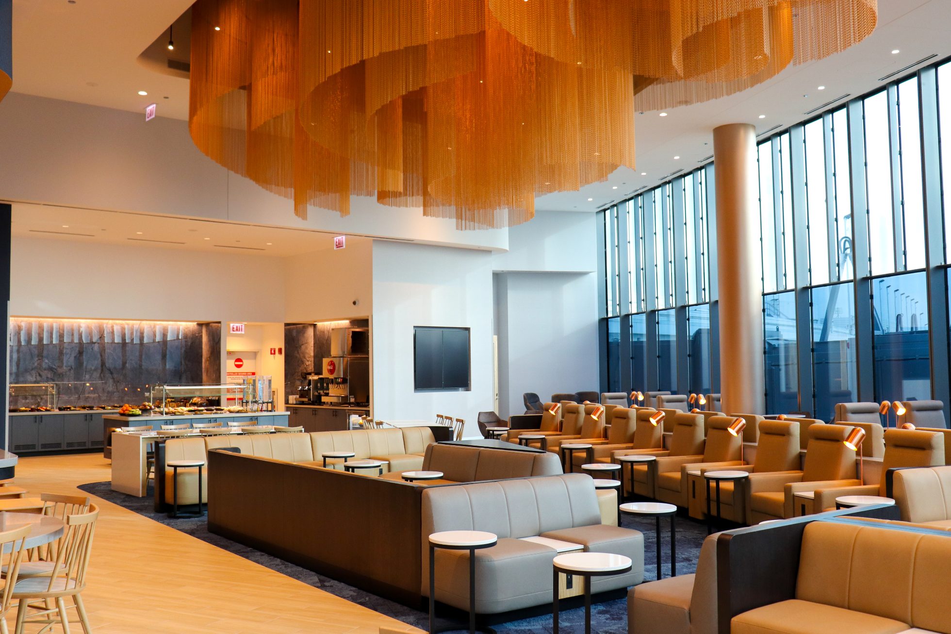 LOT Business Lounge in Chicago - reception area with illuminated LOT logo. Public space with yellow sofas, copper lamps and tables with white marble tops. Blue carpets. High ceiling and huge panoramic windows overlooking the airport. Large bar with backlights